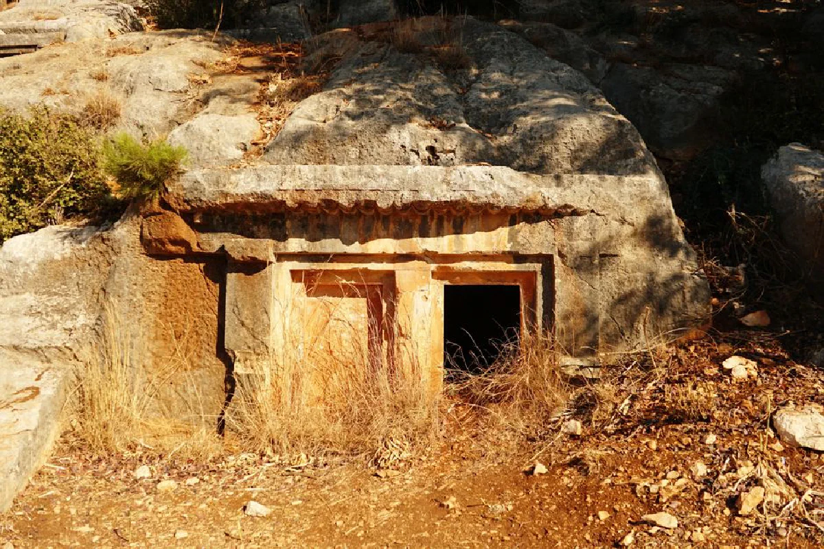 An ancient rock-cut tomb entrance hidden in a rocky, arid landscape under bright sunlight.