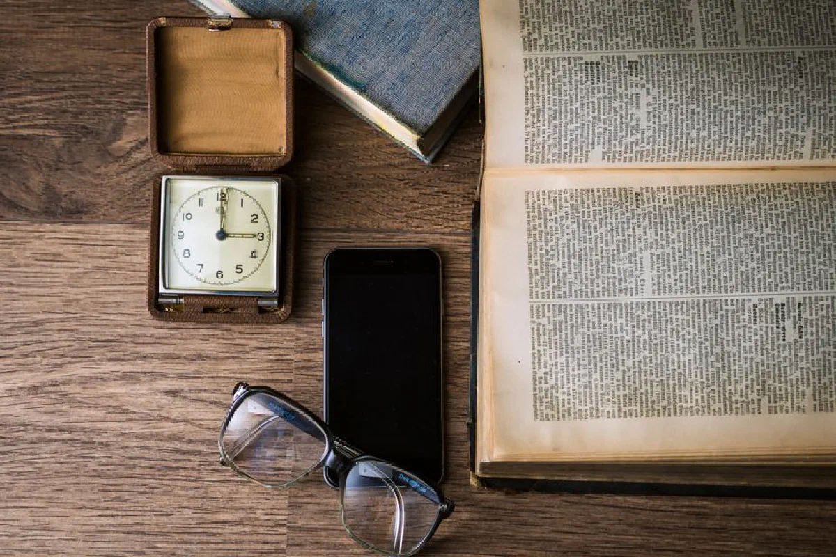 Flat lay of a vintage clock, open book, smartphone, and eyeglasses on a wooden table.