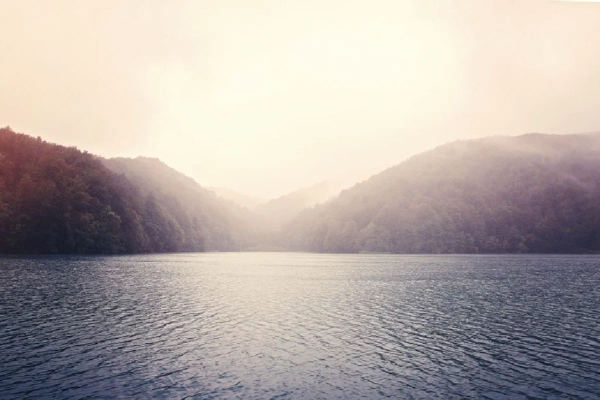 Peaceful scene of a tranquil lake surrounded by mist-covered mountains at dawn.