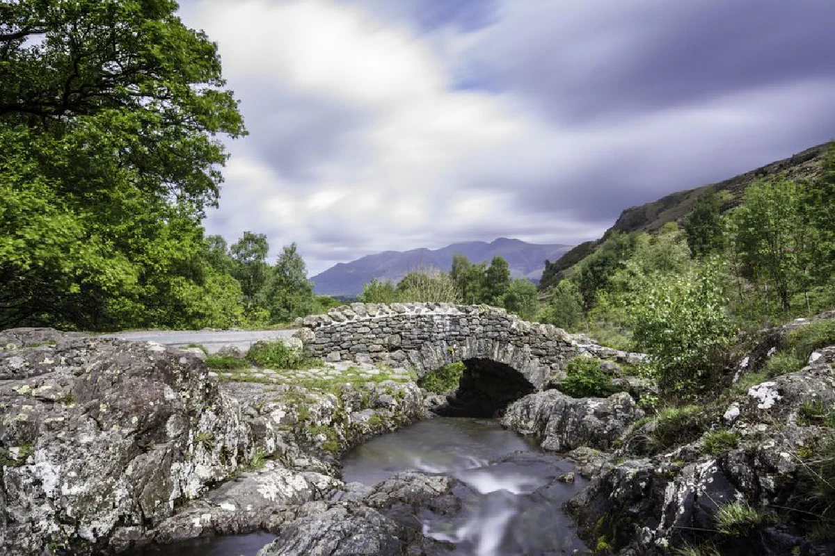 Stone bridge over a stream with lush green hills and mountains in the background.