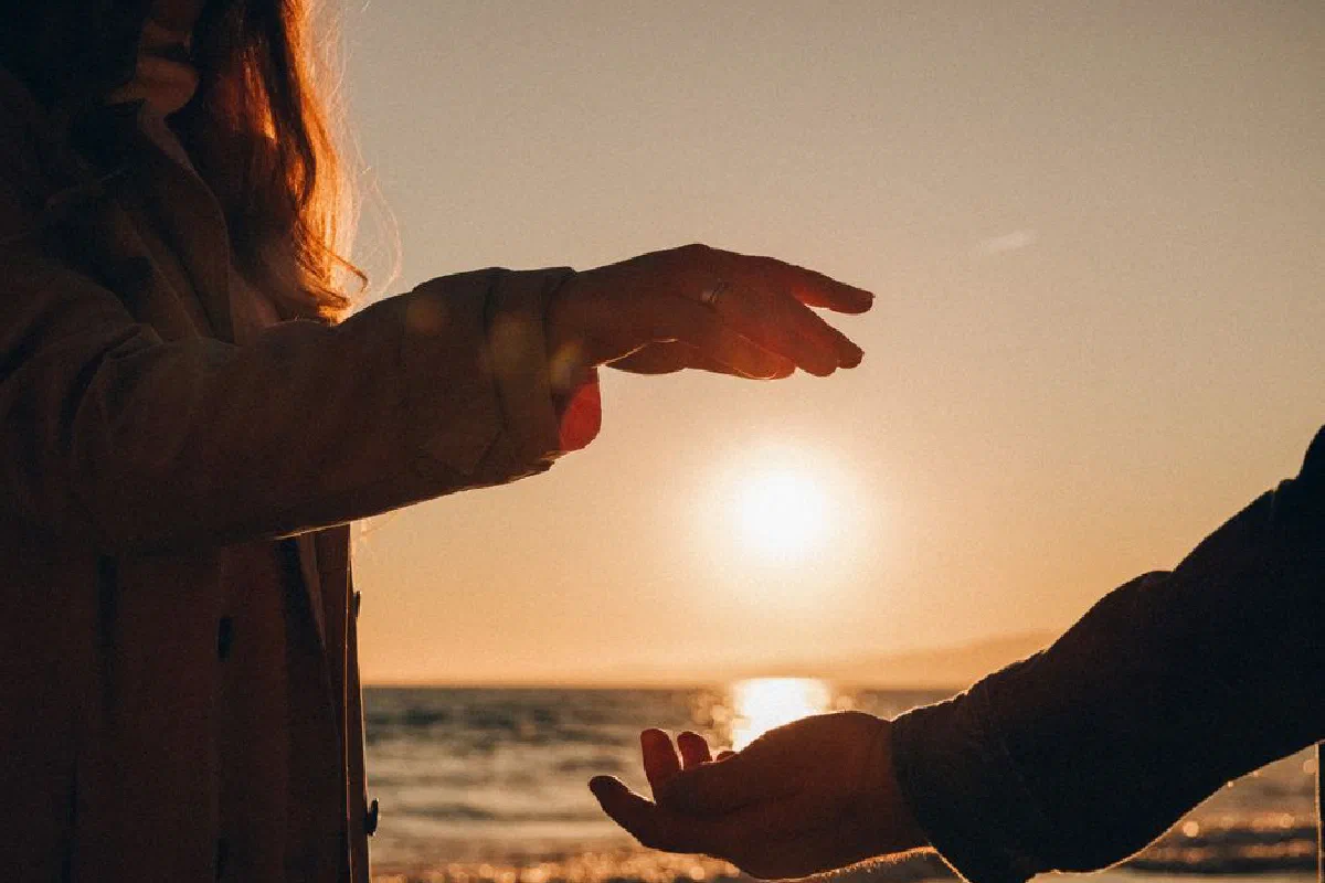 Silhouettes of a couple reaching hands at a beach during sunset, evoking romance and connection.