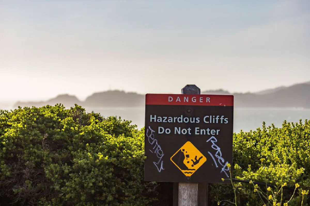 A warning sign marks hazardous cliffs by the sea amidst lush greenery.