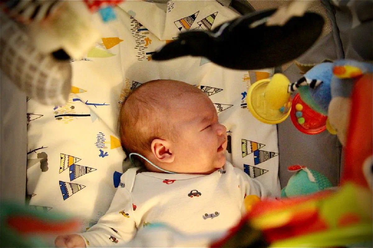 Crying newborn baby lying in a crib surrounded by colorful toys in a cozy setting.