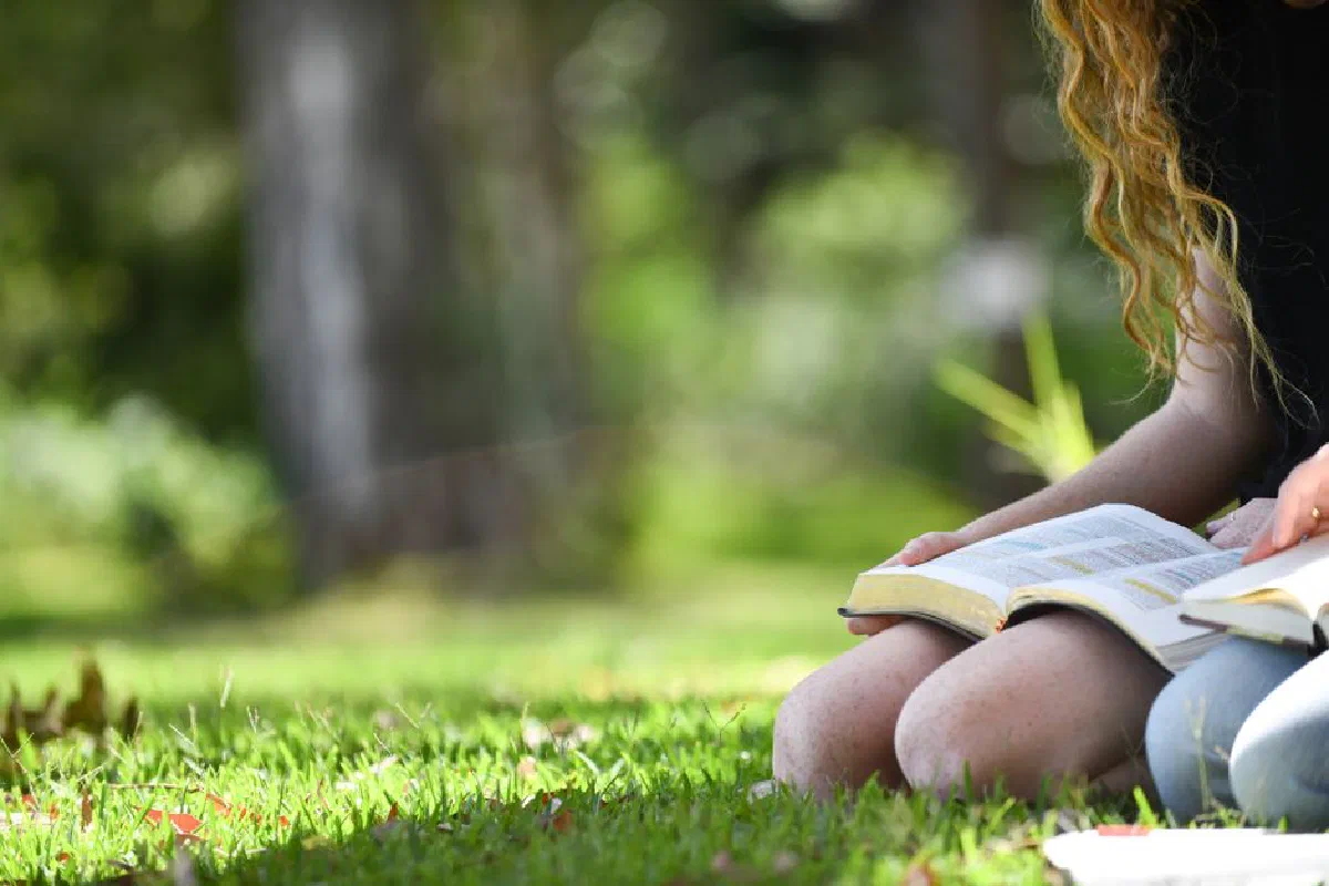 A young woman reads a book on the grass in a sunny park setting, enjoying leisure time outdoors.