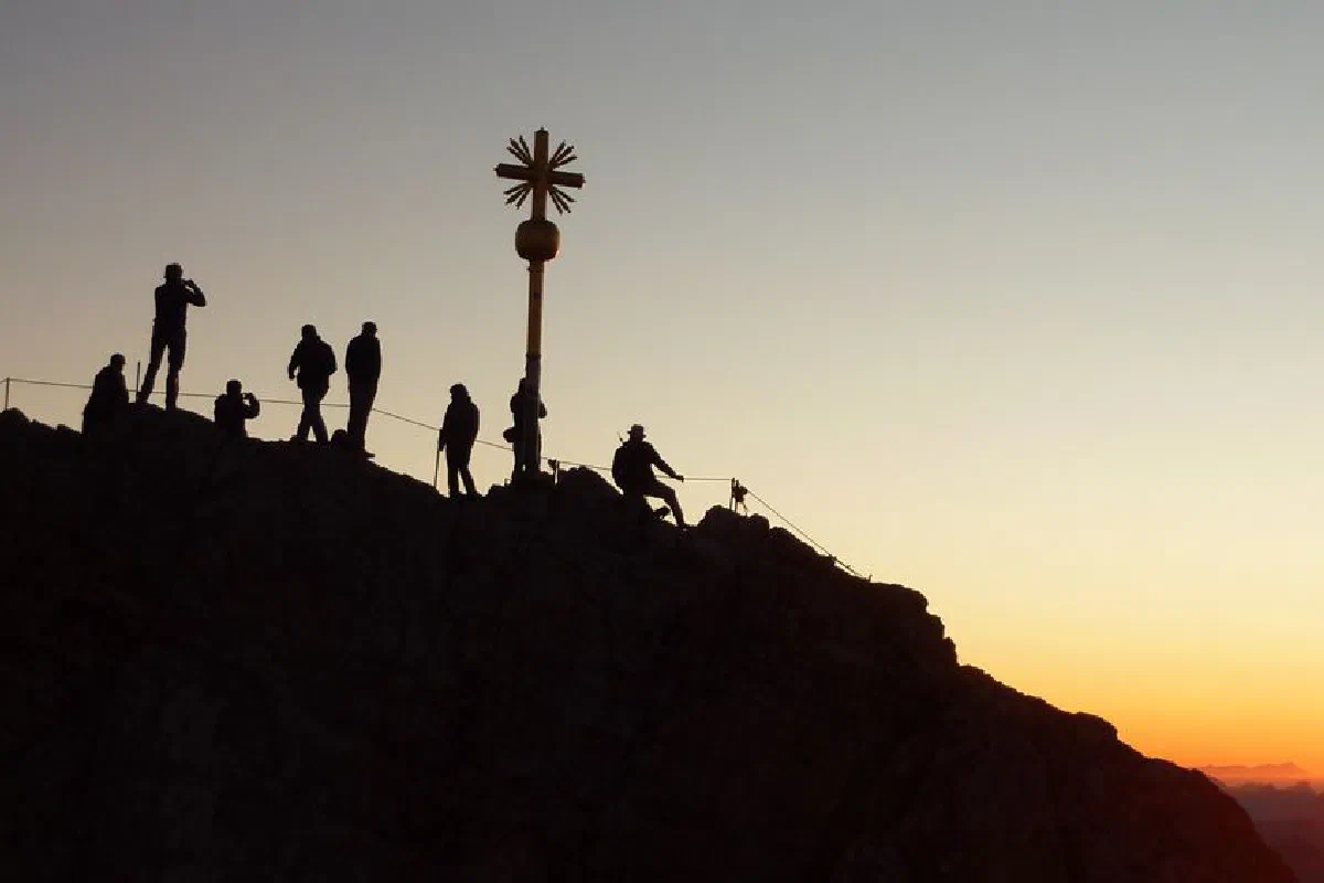 Silhouetted hikers reach a mountain summit at sunrise, with a cross marking the peak.