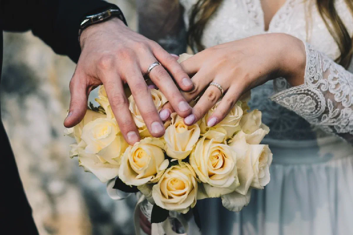 Close-up of a couple's hands displaying wedding rings above a bouquet of yellow roses.