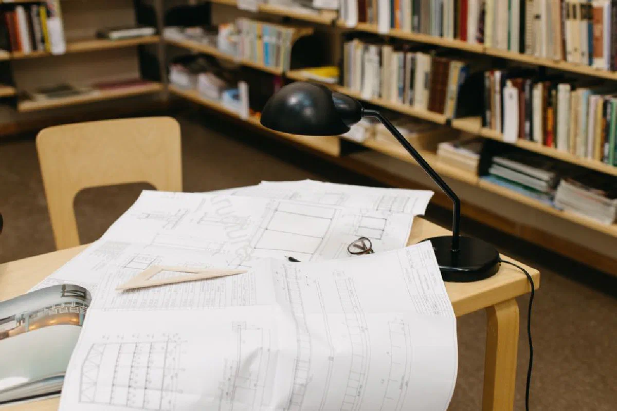 An organized collection of architectural blueprints on a desk under a lamp in a library.