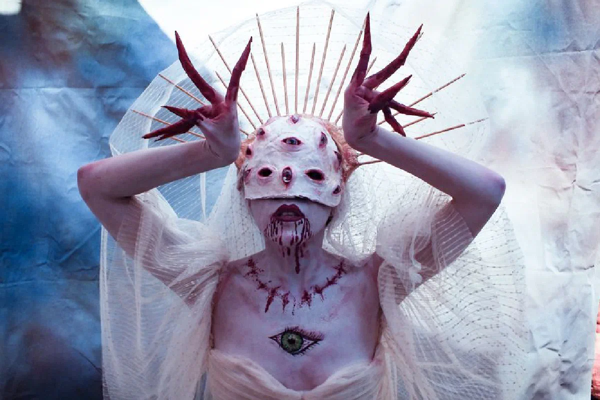 A mysterious figure in a white costume adorned with eyes, set against a dreamy backdrop.