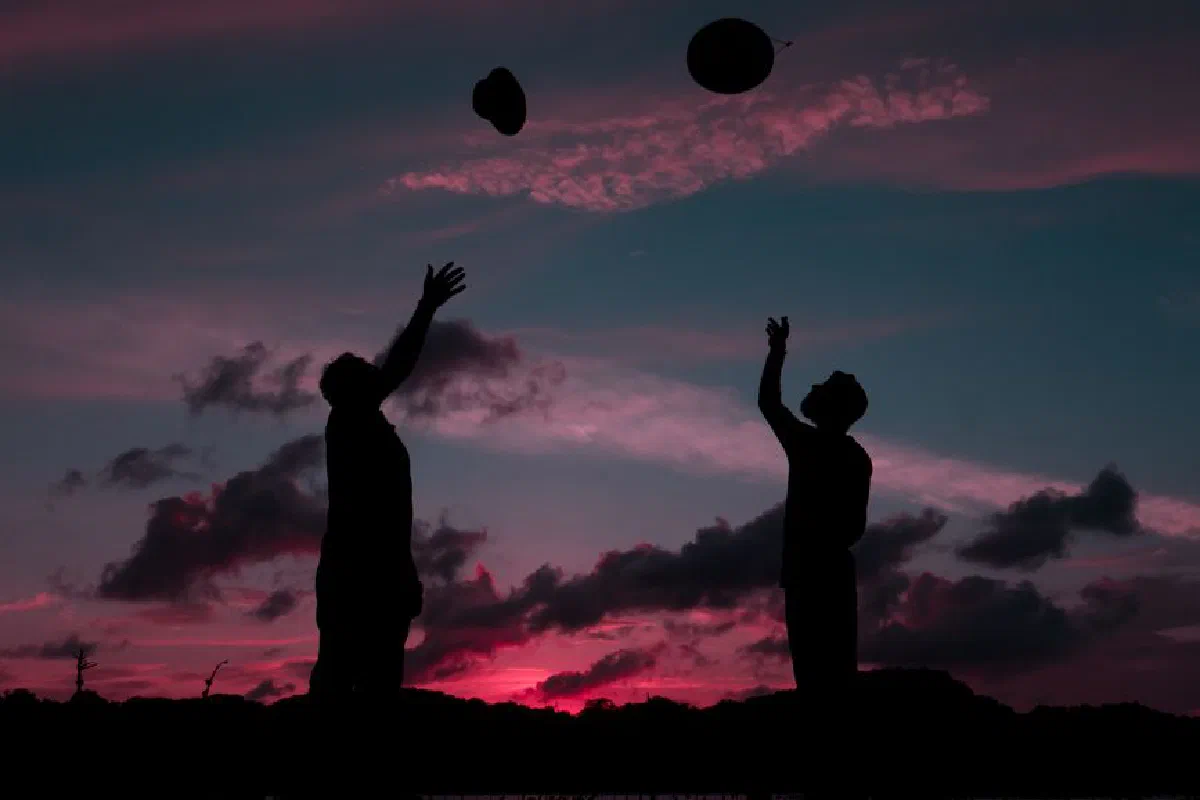 Silhouette of friends tossing hats in the air against a vibrant sunset sky.