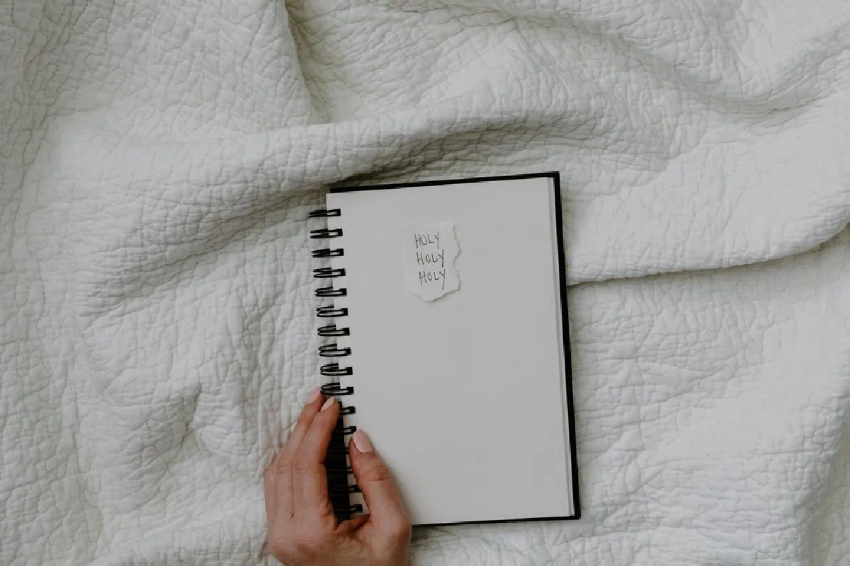 Hand holding a spiral notebook on crumpled white fabric with handwritten 'Holy Holy Holy'.