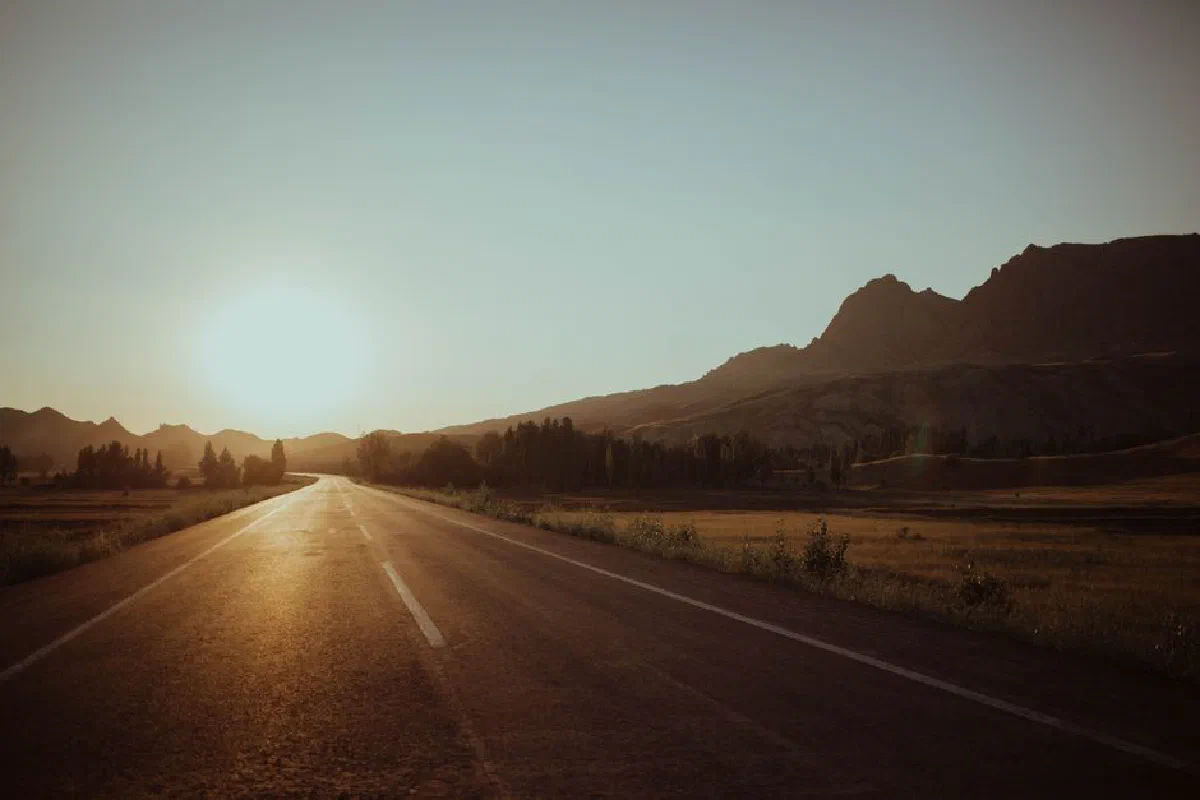 A tranquil shot of an empty highway leading into the sunset, surrounded by countryside and mountains.