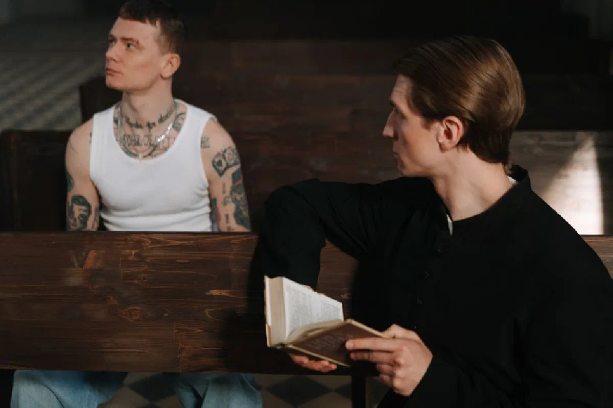 A tattooed man in a white tank top sits opposite a priest holding a holy book, symbolizing a contrast of lifestyles and beliefs.
