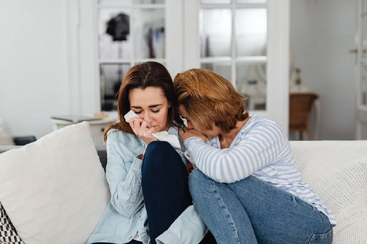 Two women sitting on a couch, comforting each other at home, displaying emotions of sadness and support.