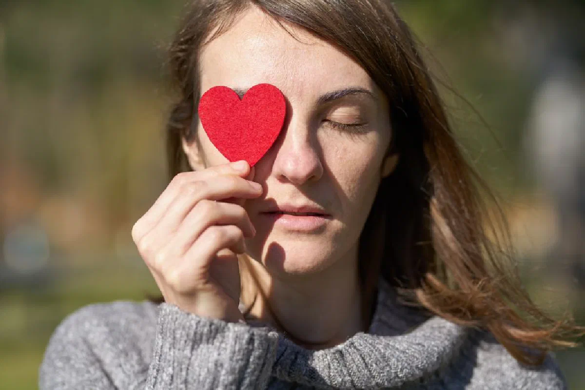 Woman holding a red heart symbol in front of her eye, enjoying a sunny outdoor day.
