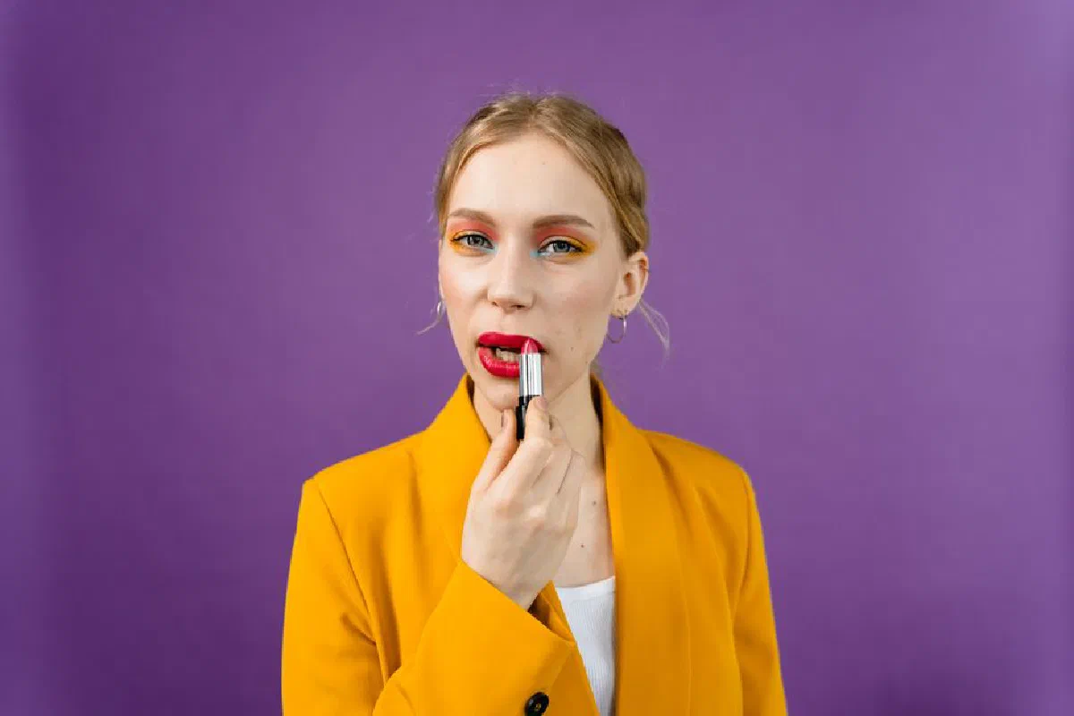 Woman in yellow blazer applying bright red lipstick with clear purple backdrop.