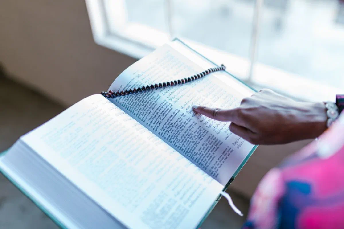 A Muslim woman pointing to a passage in the Quran with prayer beads draped over it indoors.