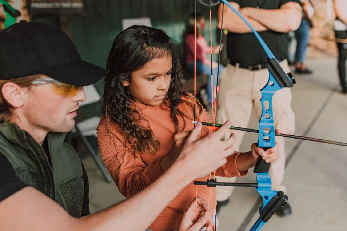 Young girl learning archery from an instructor at an outdoor range.