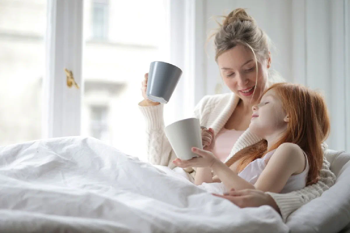 A mother and daughter sharing a warm moment with cups in bed, enjoying a cozy morning together.