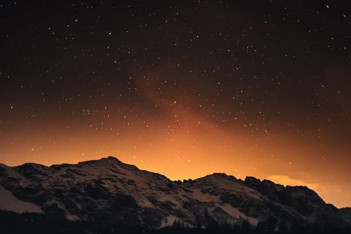 Captivating view of starry night sky over snow-covered mountains in Italy with a warm, orange glow.