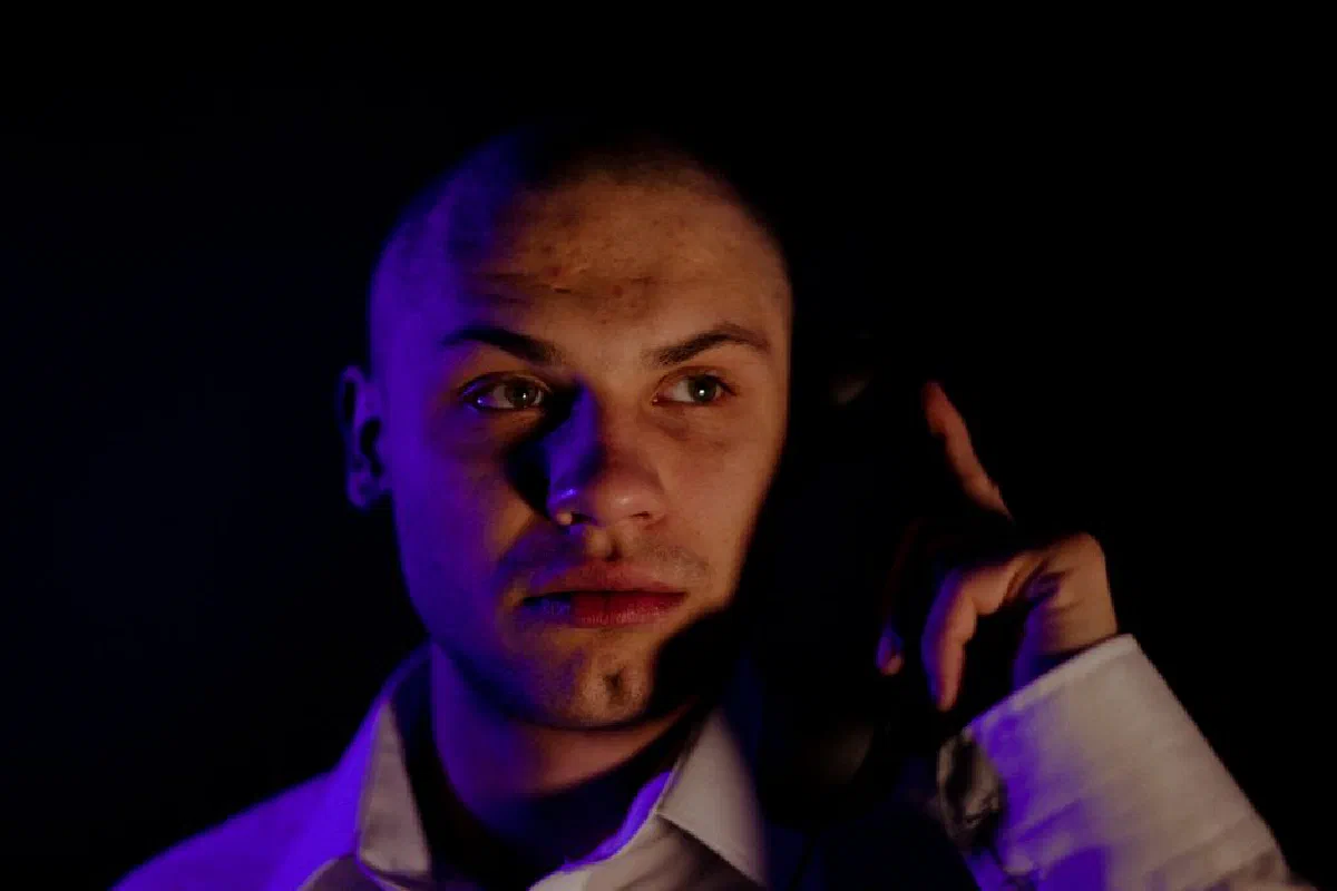 Close-up of a bald man in white shirt holding a telephone in a dramatic, moody setting.