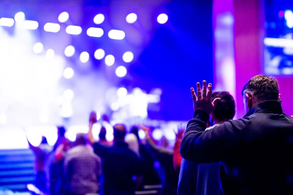 A lively and engaging indoor worship celebration filled with a diverse crowd immersed in prayer and music under bright stage lights.