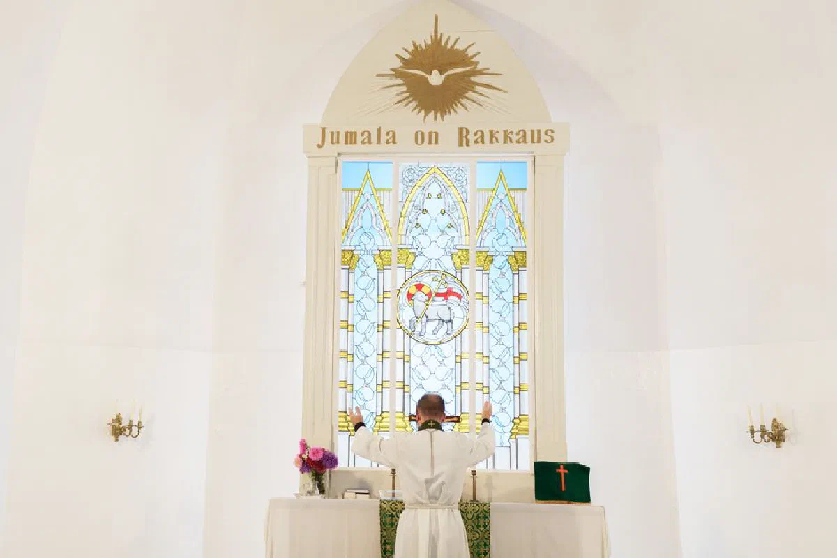 A pastor stands at an altar, leading a prayer in a beautifully lit church interior with stained glass windows.