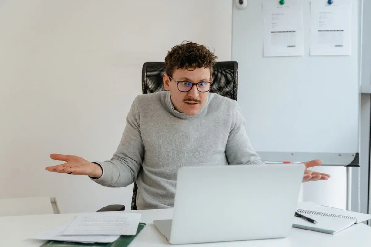 A man in a gray sweater and glasses gesturing at his laptop screen during a video call in an office setting.