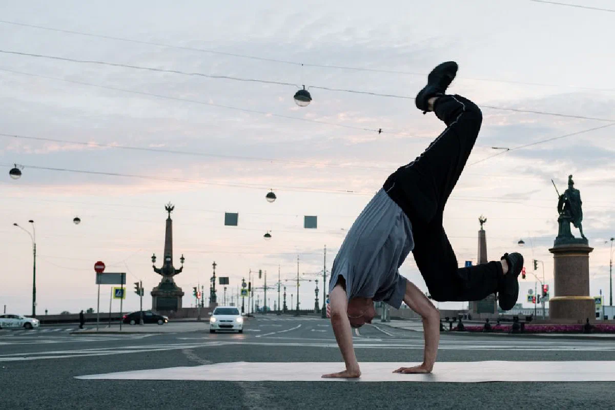A man performing a handstand in an urban street setting with statues in the background at dusk.