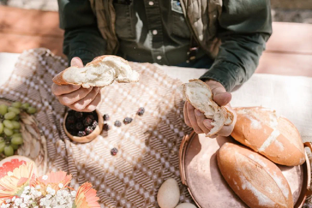A person at an outdoor picnic holding fresh bread with seasonal fruits and flowers.