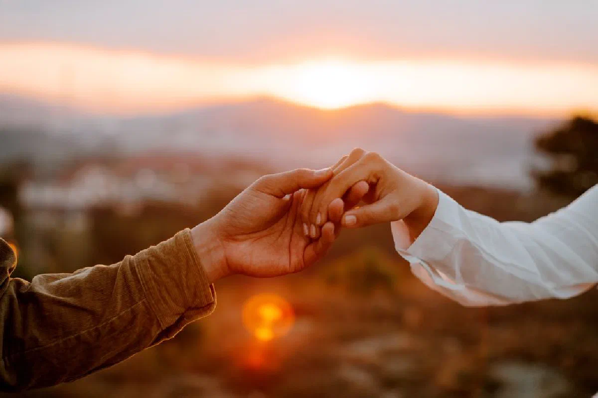 Crop faceless couple holding hands against picturesque mountains under colorful sky at sundown