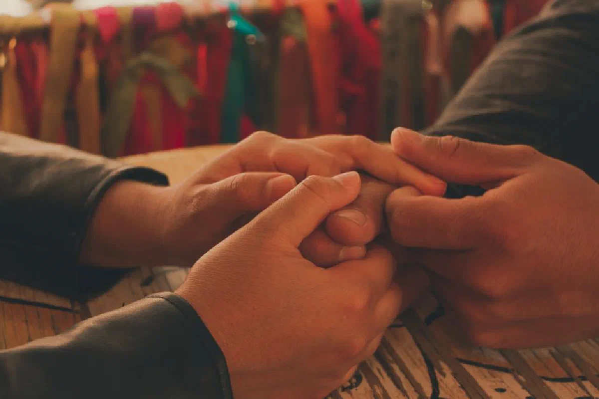 Warm and intimate close-up of a couple holding hands on a wooden table with colorful ribbons in background.