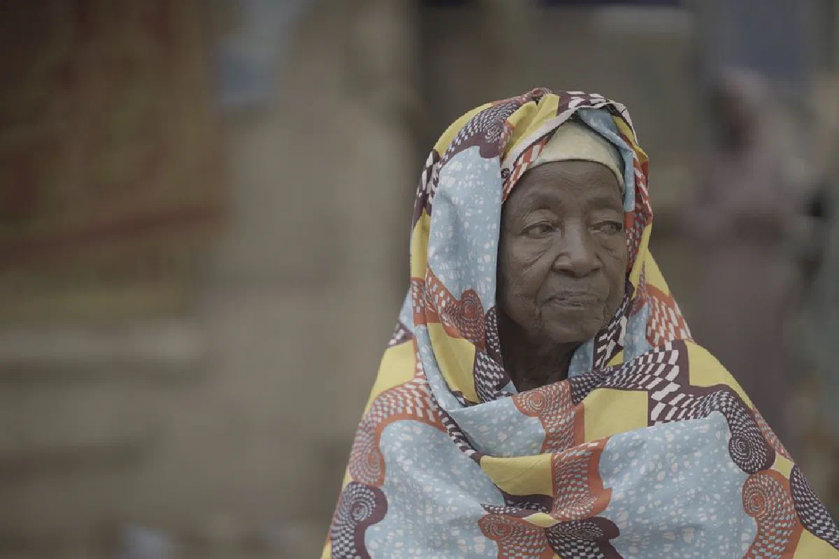 An elderly woman wearing a colorful Islamic dress stands outdoors in Ghana.