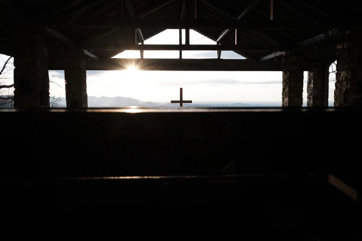 A serene view of a chapel cross silhouetted against a sunlit mountain landscape.