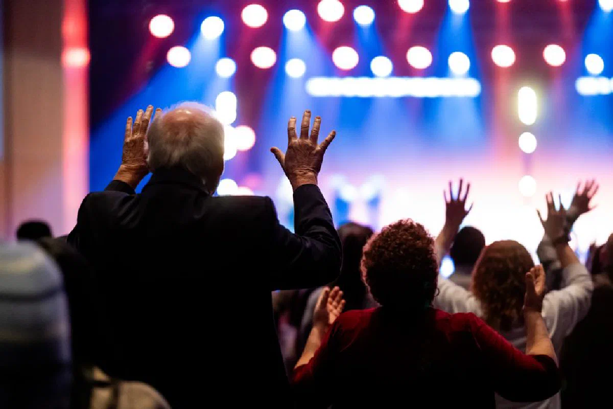 Dynamic scene of a worship concert with people joyfully raising their hands in praise under colorful stage lights.