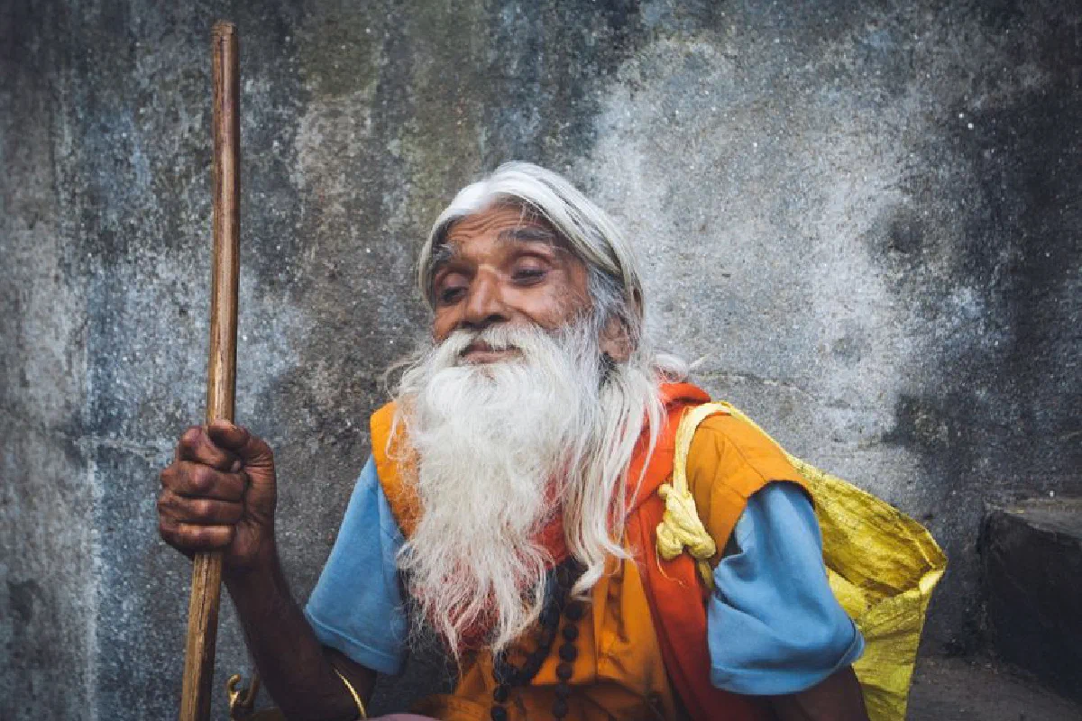 Portrait of an elderly man with a long beard sitting outdoors against a textured wall, holding a stick.