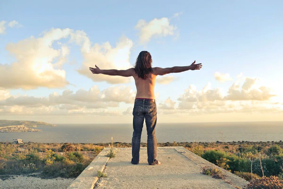 A man embraces freedom with arms wide open, enjoying sunset over Mgarr, Malta coast.