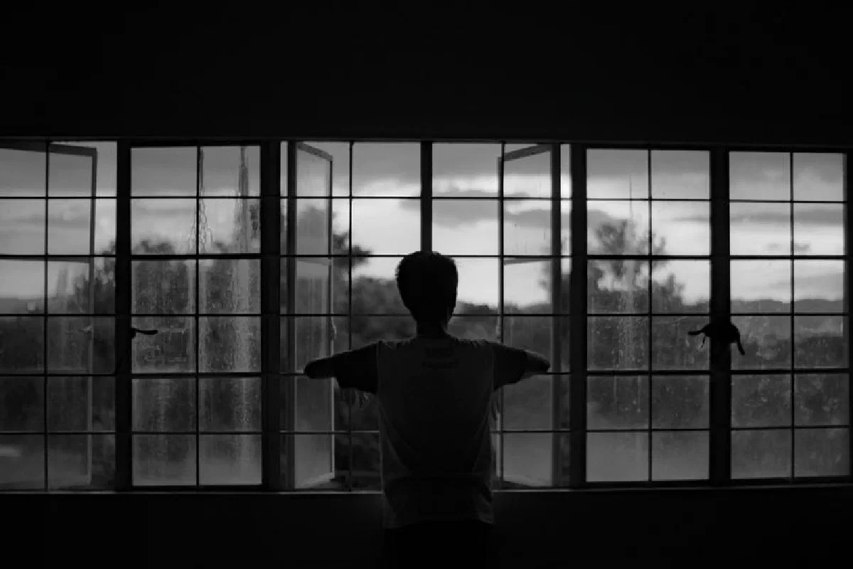 A moody silhouette of a man staring out an open window, evoking solitude.