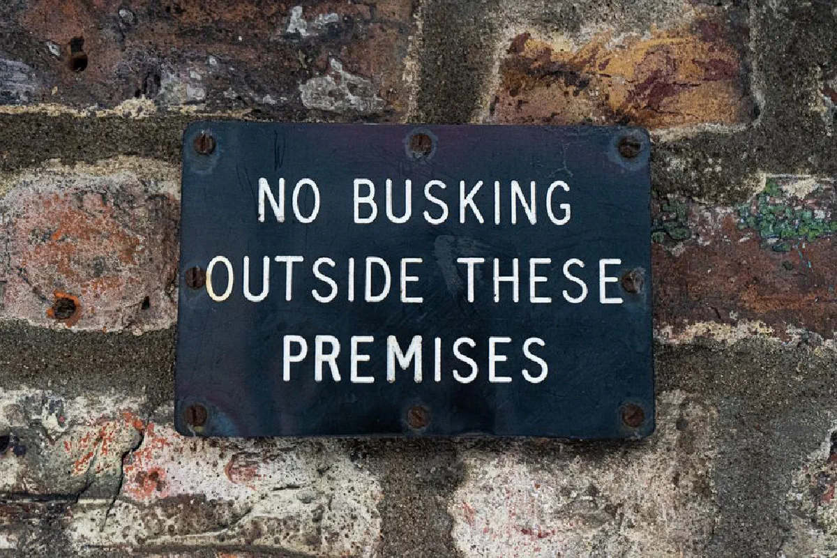 Black sign stating 'No Busking' affixed to a textured brick wall surface.