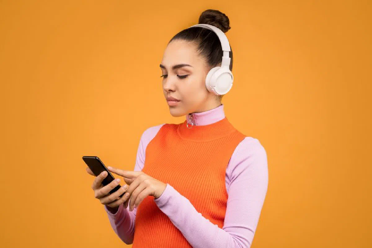 Woman with white headphones using smartphone against an orange background.