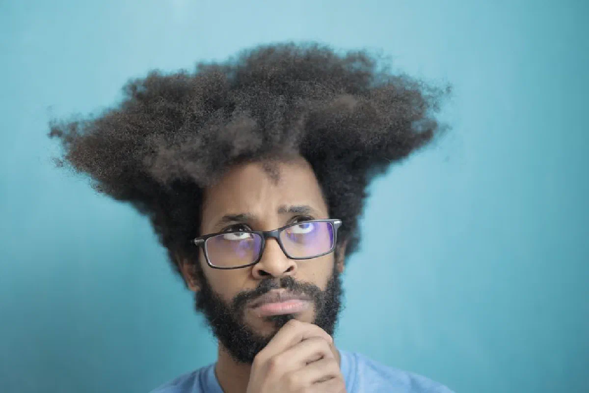 Portrait of a thoughtful man with a unique afro hairstyle, wearing glasses against a blue backdrop.