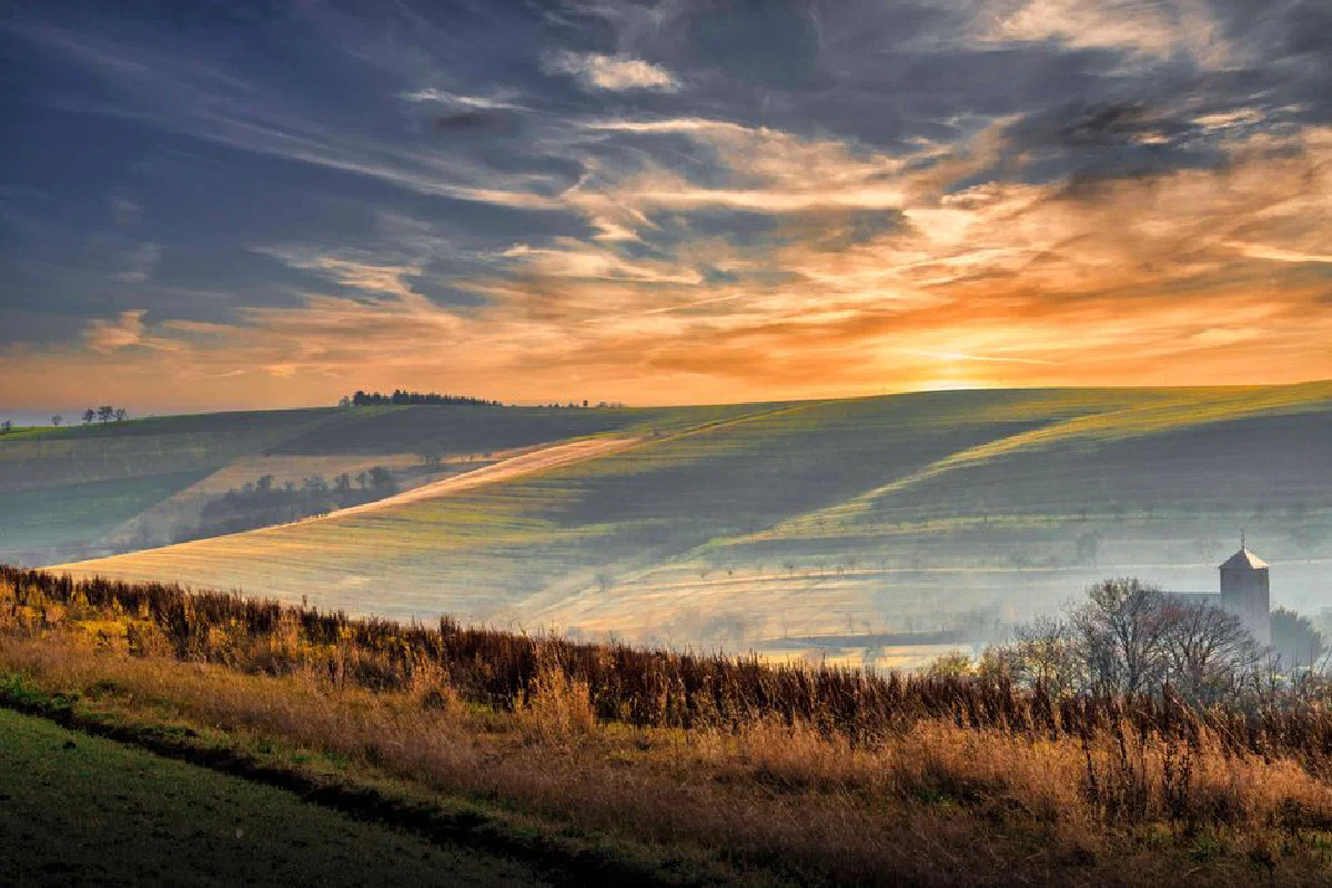 Breathtaking sunrise over rolling hills with fields and rural landscape, featuring mist and vibrant colors.