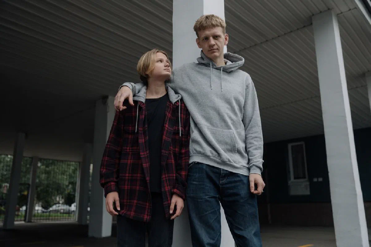 Two brothers in casual attire, standing under a shelter, showcase sibling bond and casual fashion.