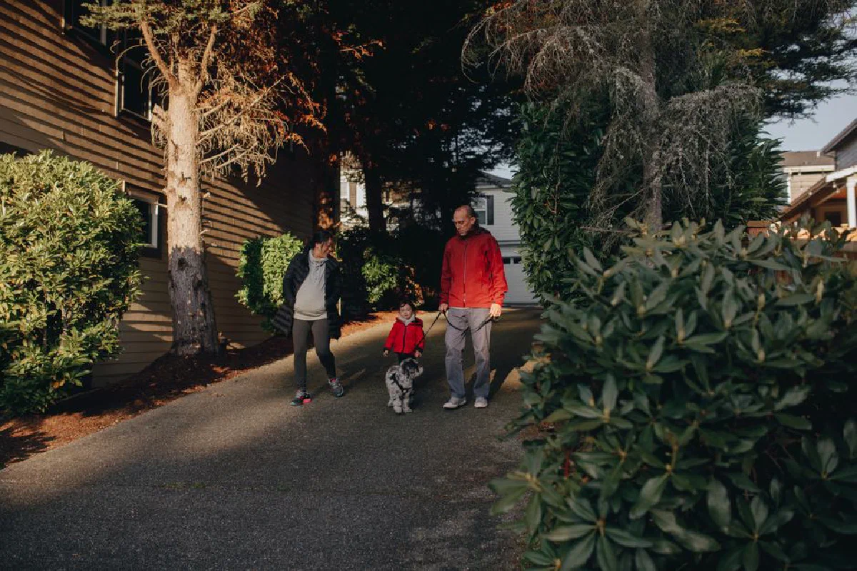 A family enjoys a walk with their pet dog on a sunny day, surrounded by lush greenery in a suburban neighborhood.