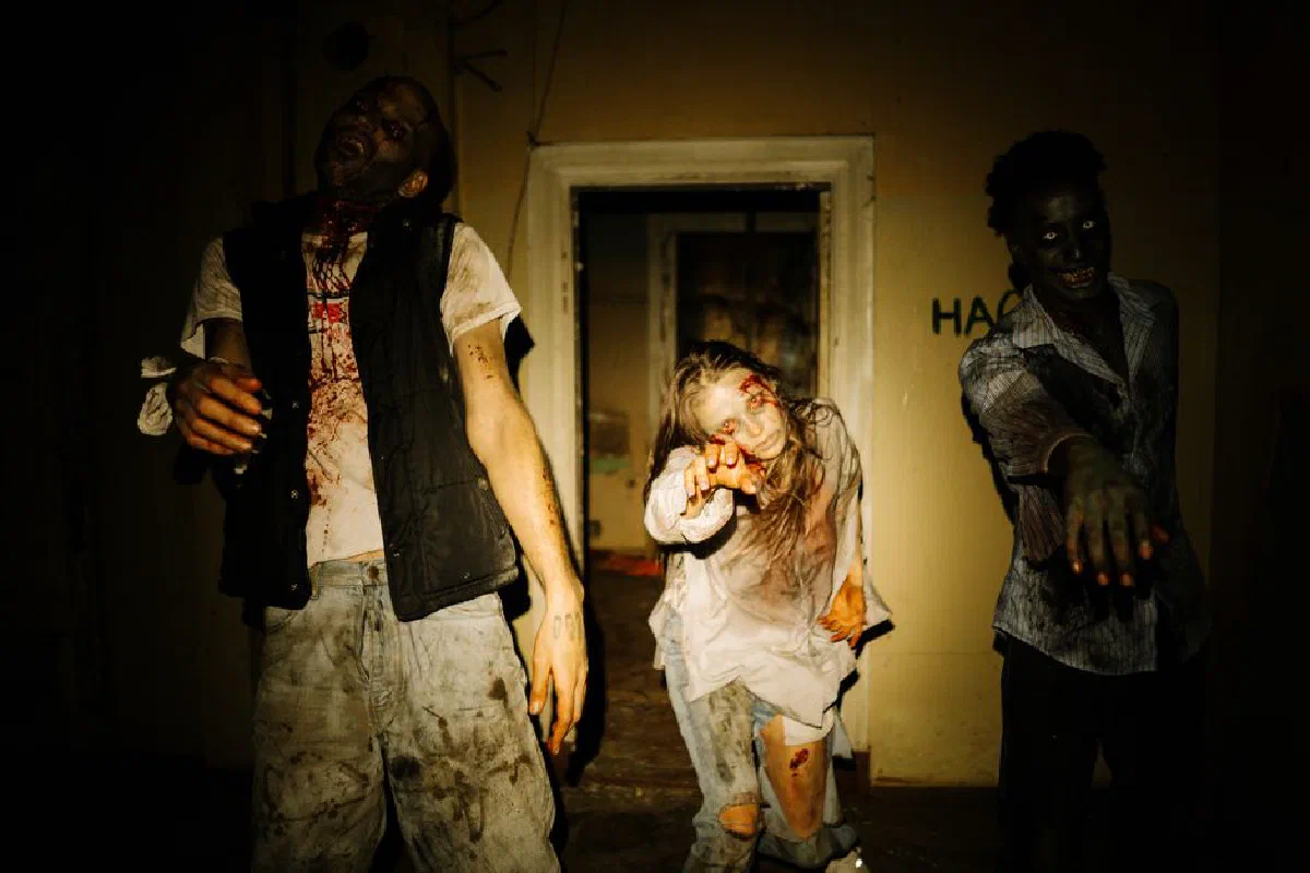 Three costumed zombies outstretch their hands in a dimly lit, abandoned house setting.