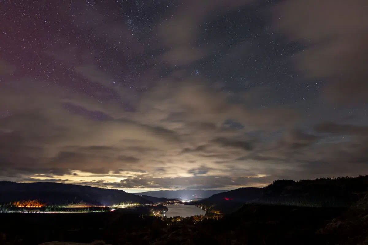 A dramatic night sky filled with stars above a peaceful lake, enveloped by distant hills and glowing city lights.