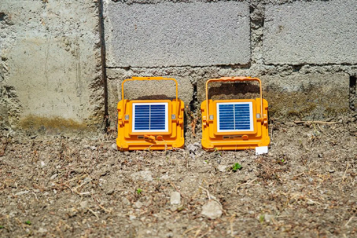 Two orange portable solar lamps placed on the ground against a concrete wall, absorbing sunlight.