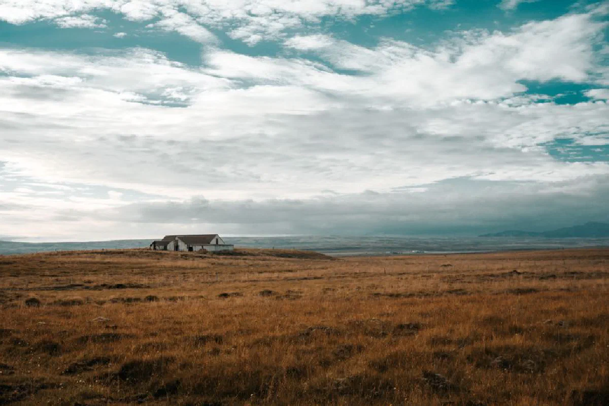 A solitary abandoned house stands amidst expansive rural grasslands under a cloudy blue sky.