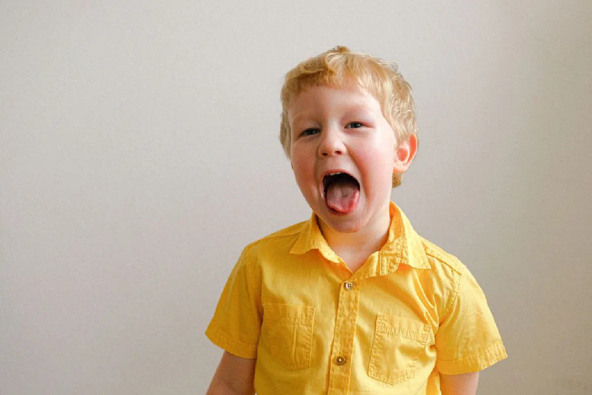 A cute child in a bright yellow shirt playfully sticking out their tongue against a neutral background.
