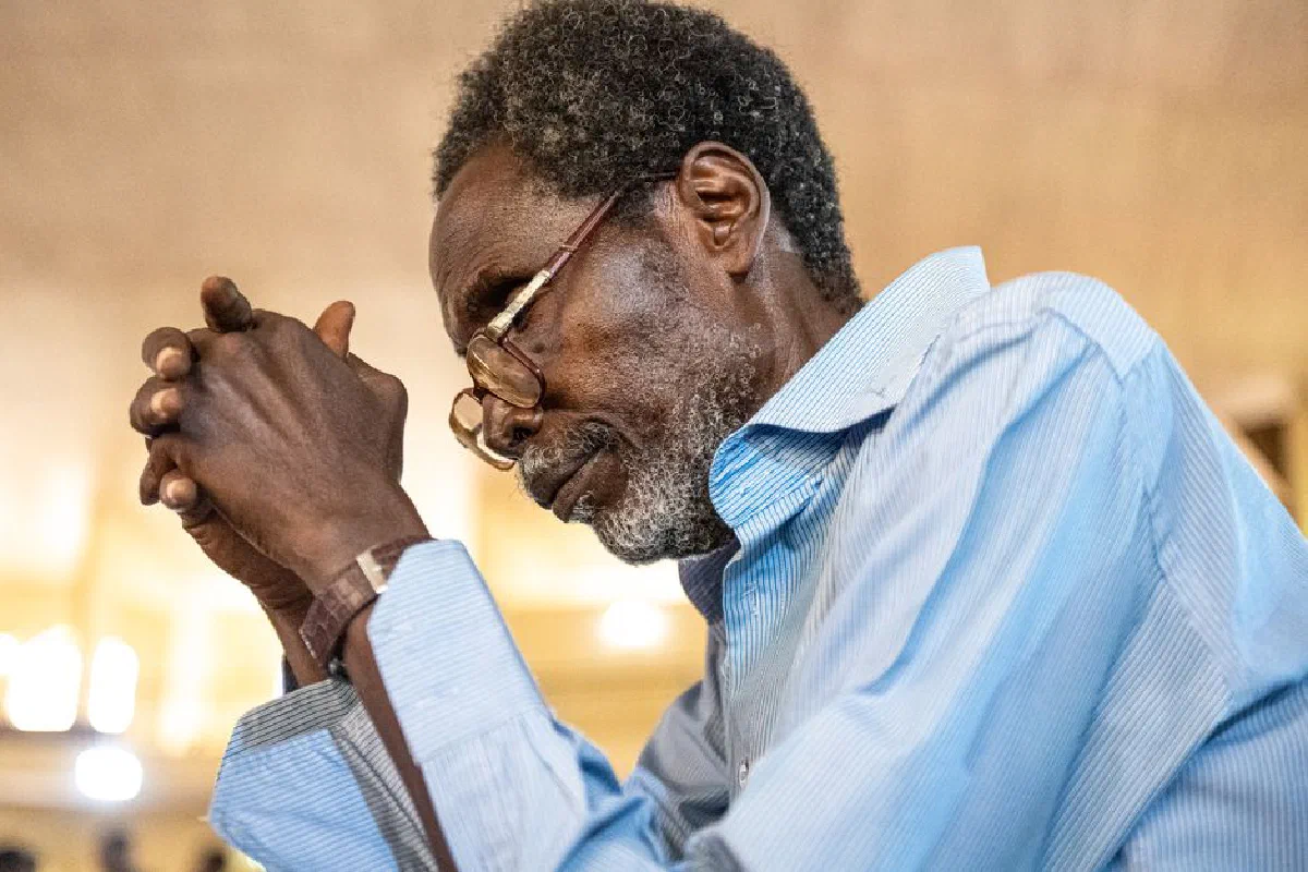 A senior man in a blue shirt, wearing glasses, praying with hands clasped indoors.