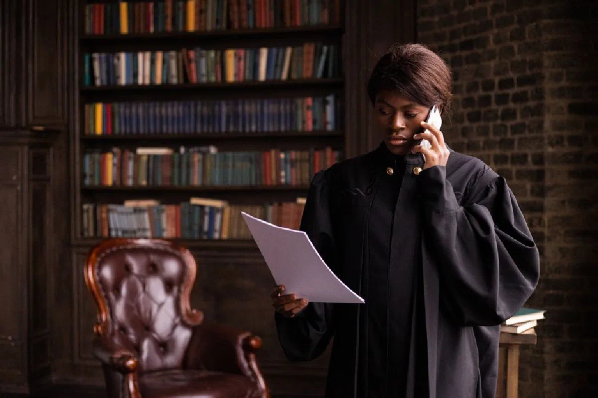 African American judge in traditional robes holds a document and talks on the phone in a law library.